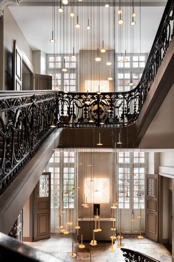 Suspensions lumineuses entre les étages de l’escalier, rampes en ferronnerie et hautes fenêtres à La Cour des Consuls.
