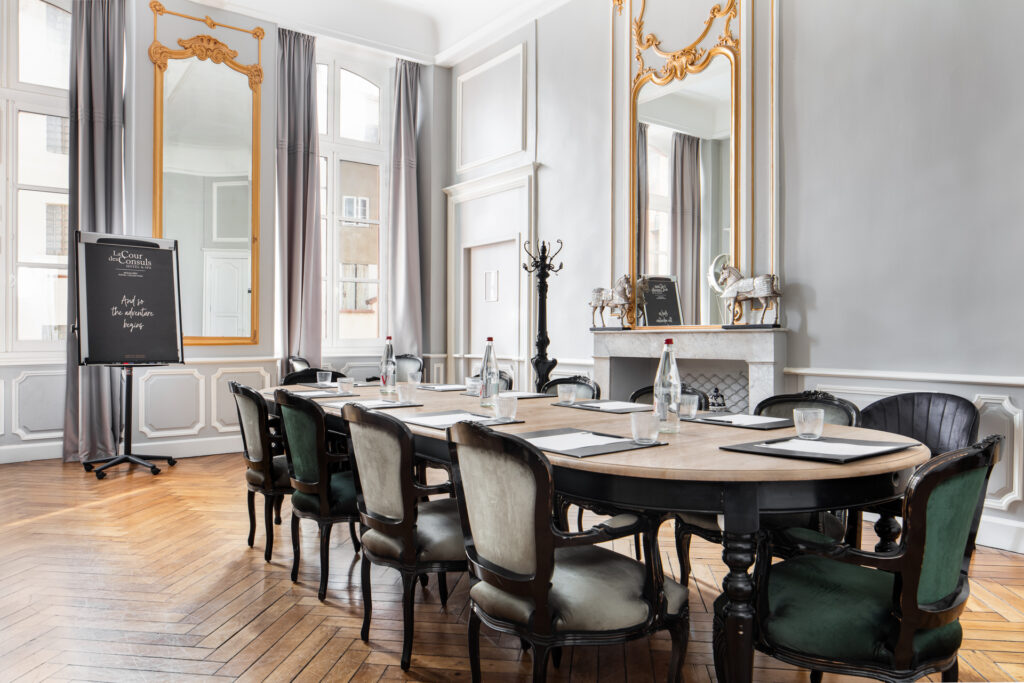 Grande table de réunion entourée de chaises, entre de hautes fenêtres, des miroirs dorés et une cheminée.