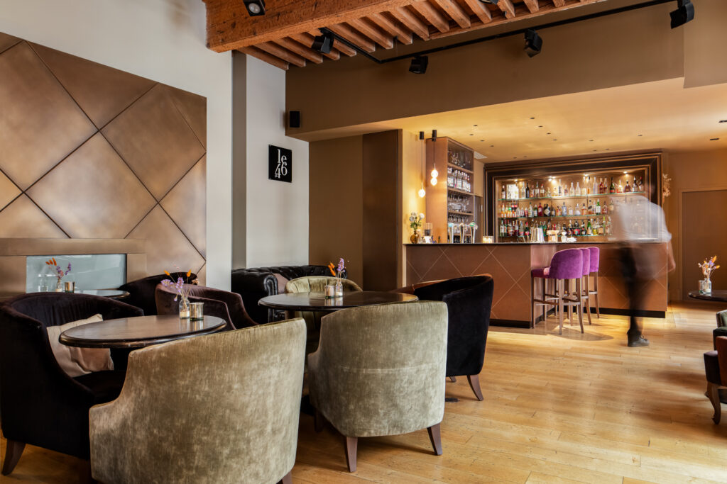 Bar Le 46 avec fauteuils en velours, poutres apparentes et silhouette en mouvement devant le comptoir.