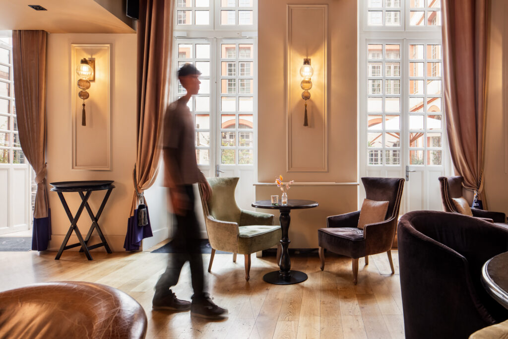 Silhouette en mouvement dans le salon de La Cour des Consuls, devant les fauteuils et les hautes fenêtres sur cour.