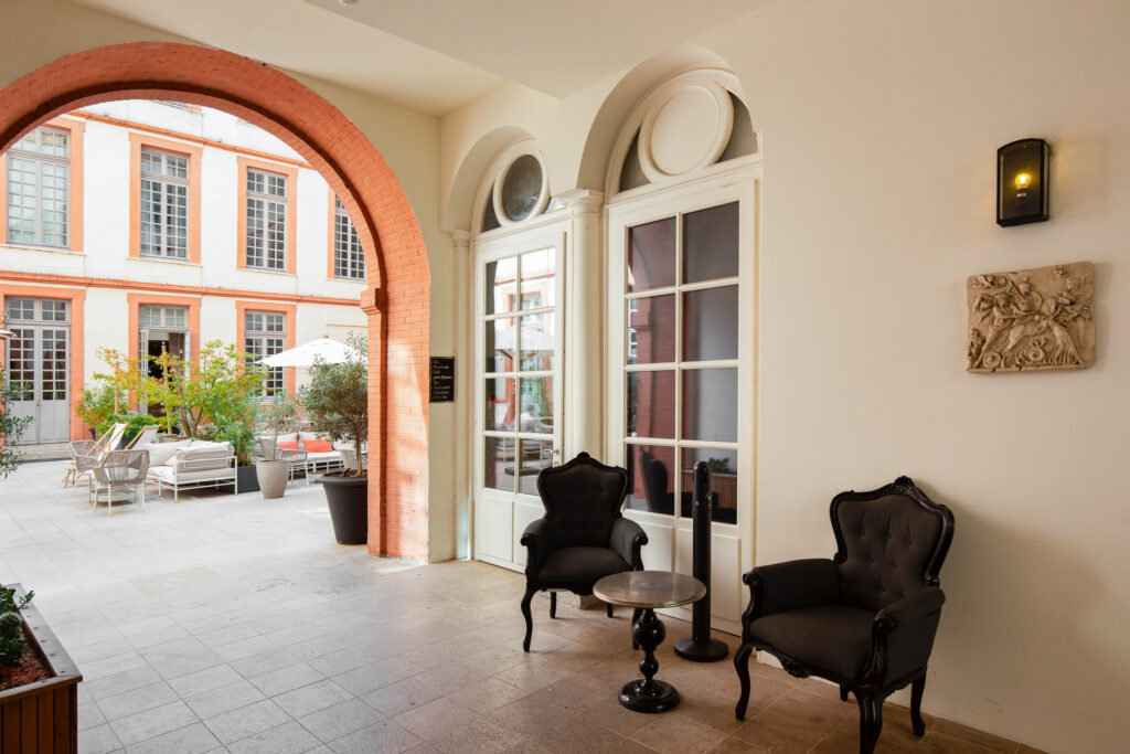 Passage voûté ouvrant sur la cour intérieure, avec deux fauteuils noirs et une petite table le long du mur.