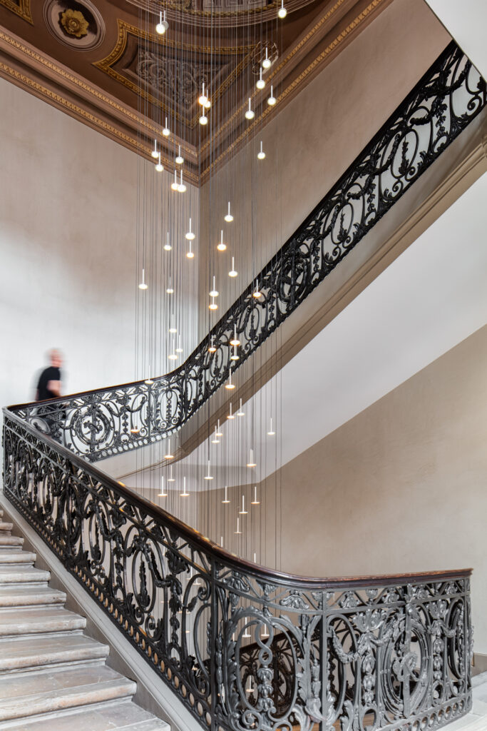 Vue ascendante de l’escalier aux rampes ouvragées, avec une silhouette sur le palier et un plafond peint.