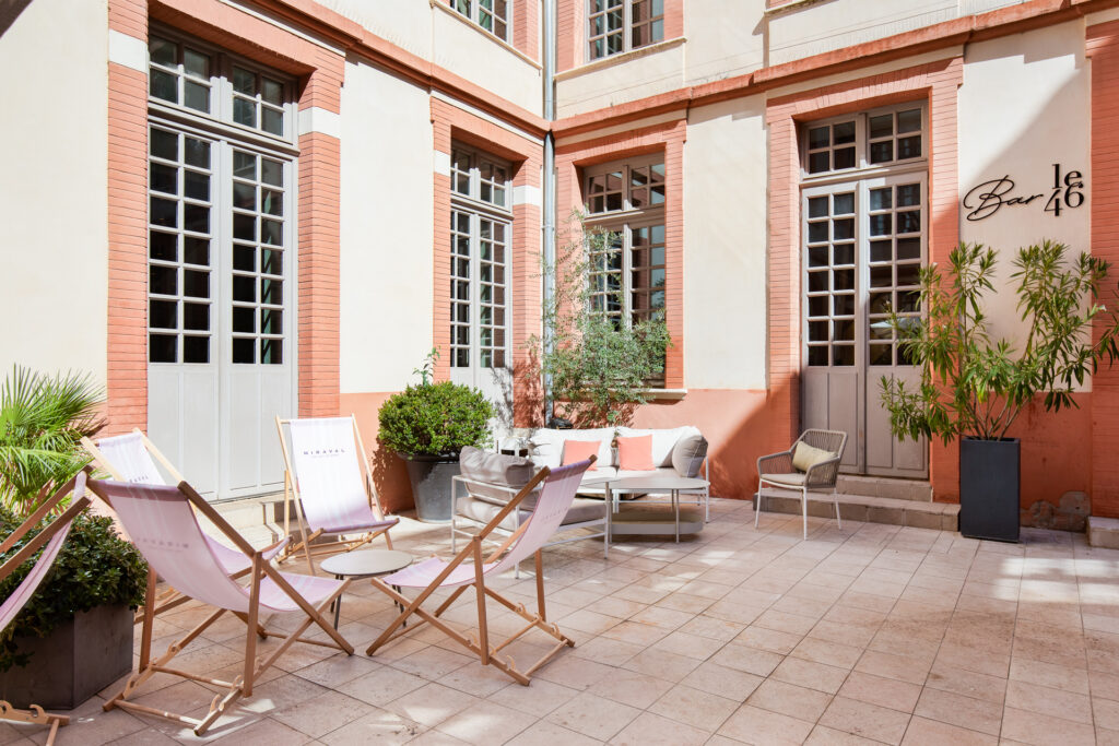 Terrasse avec chiliennes, fauteuils et plantes en pots, entourée de façades claires aux encadrements de brique rose.