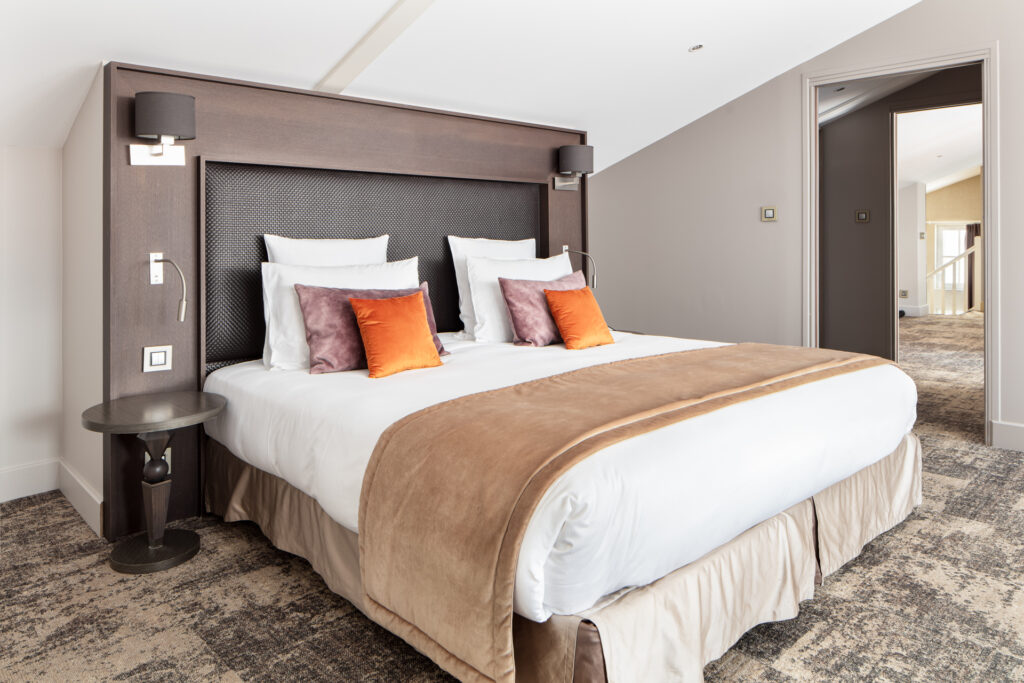 Suite avec grand lit, tête de lit brune et coussins orangés, ouverte sur le salon attenant.