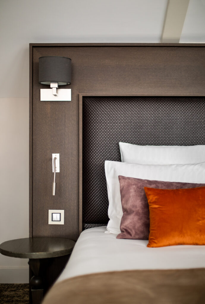 Détail d’une tête de lit brune, d’une applique et de coussins mauve et orange à La Cour des Consuls.