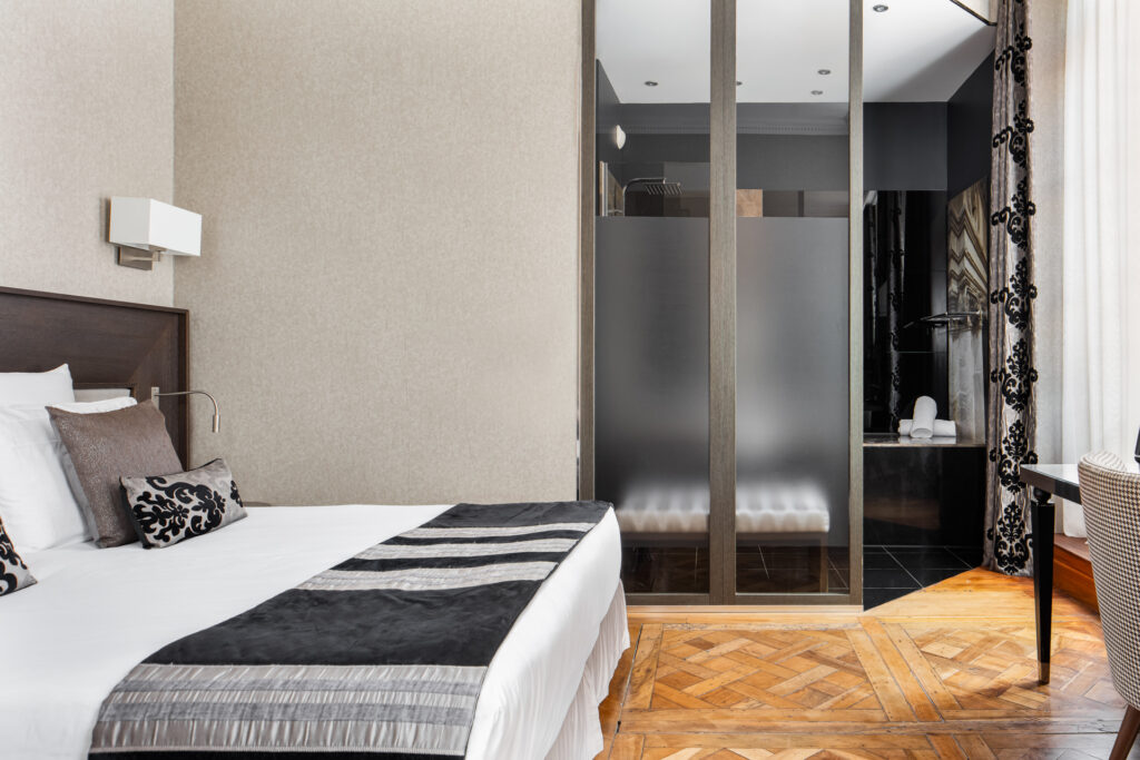 Chambre au linge de lit noir et blanc, avec parquet à chevrons et ouverture vitrée vers la salle de bains.