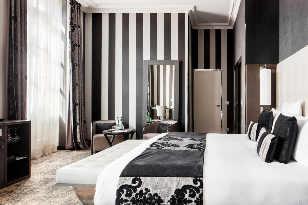 Chambre aux murs rayés gris et blanc, avec grand lit, banquette et coin salon près d’une haute fenêtre.