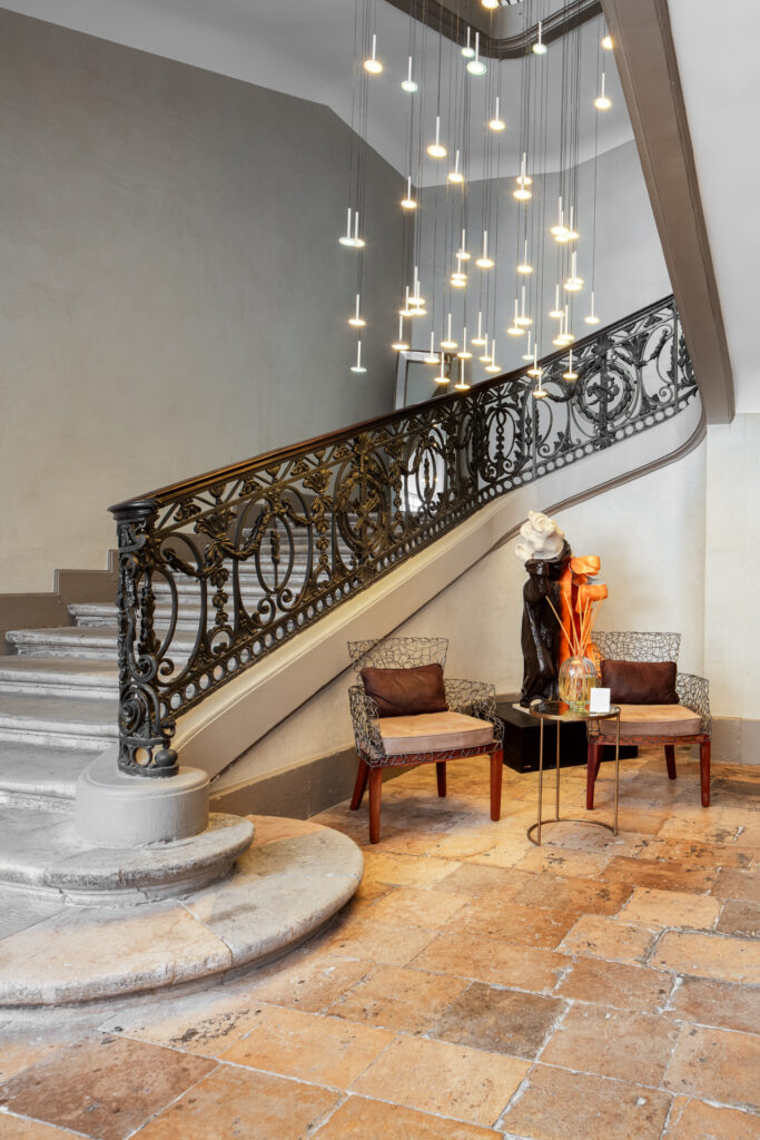 Départ de l’escalier en pierre, rampe ouvragée et deux fauteuils autour d’une sculpture à La Cour des Consuls.