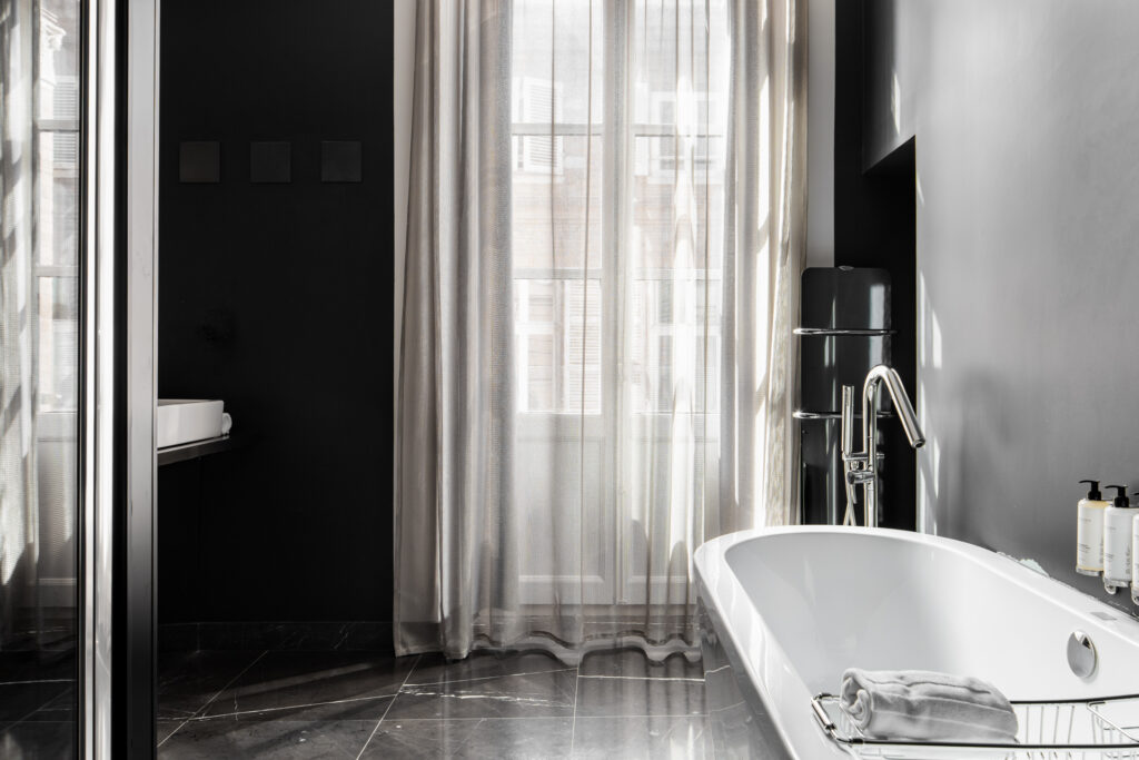 Baignoire îlot blanche dans une salle de bains sombre, près d’une haute fenêtre habillée de voilages.