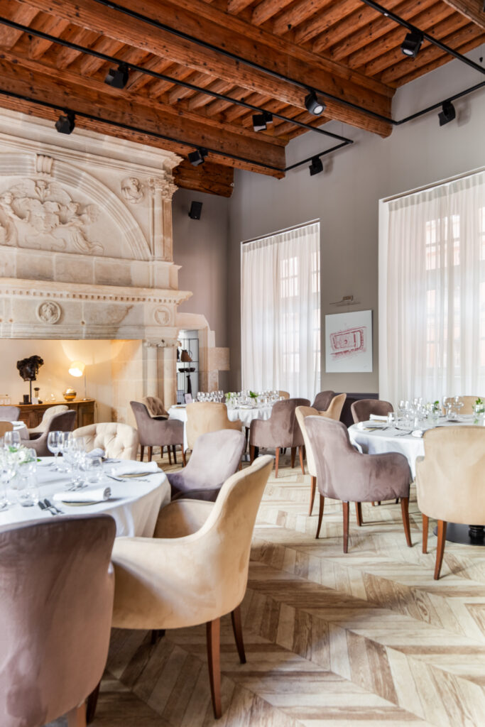 Vue du restaurant avec plafond à poutres apparentes, cheminée sculptée, hautes fenêtres et parquet à chevrons.
