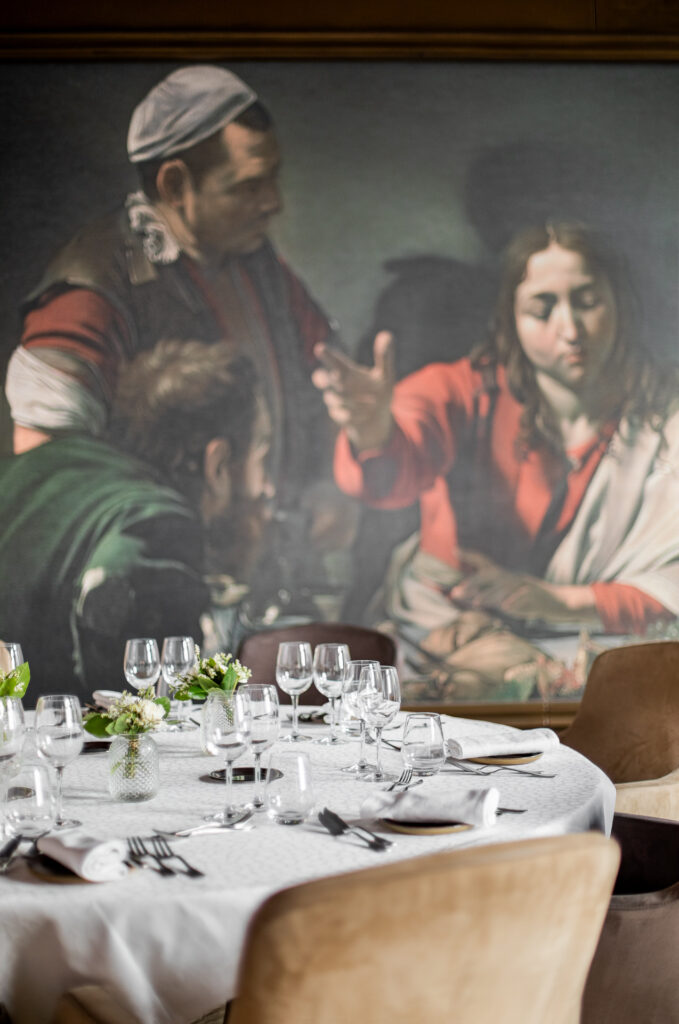 Table ronde dressée, verres et fauteuils en velours beige devant un tableau de Caravage au restaurant.
