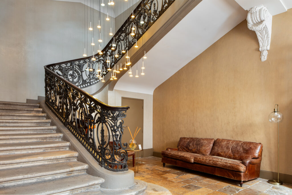 Escalier en pierre et canapé en cuir brun dans le hall de La Cour des Consuls, sous les suspensions lumineuses.