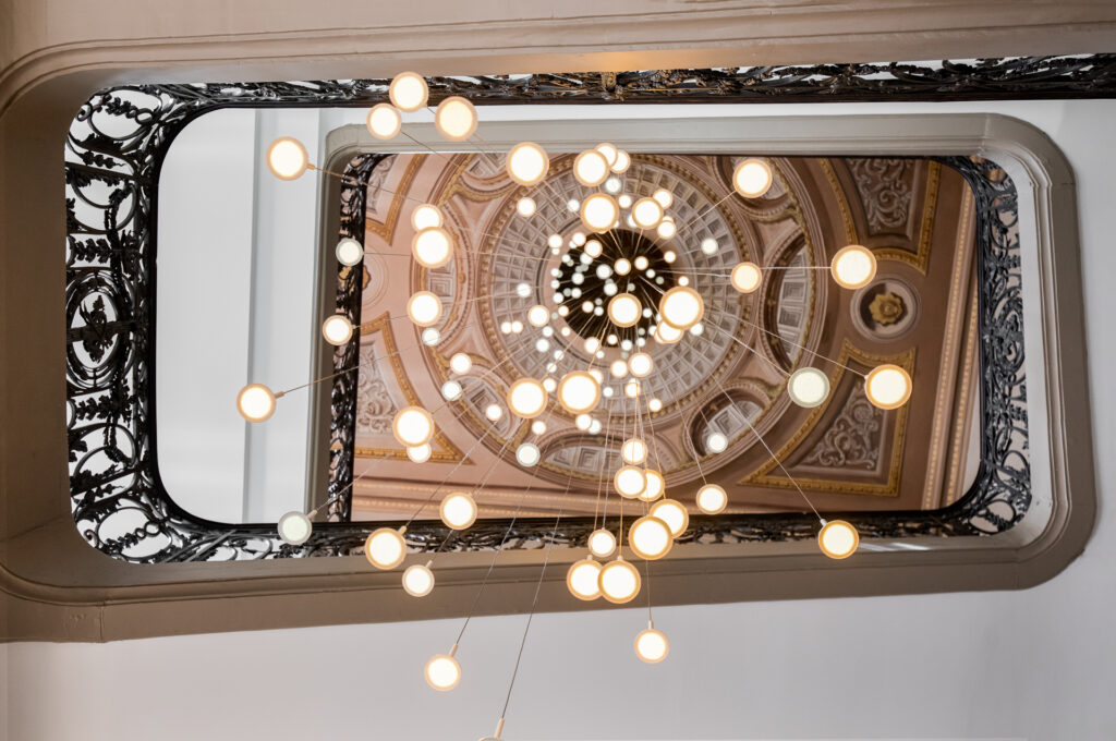 Vue verticale vers le plafond peint de la cage d’escalier, encadré par les ferronneries et les suspensions lumineuses.