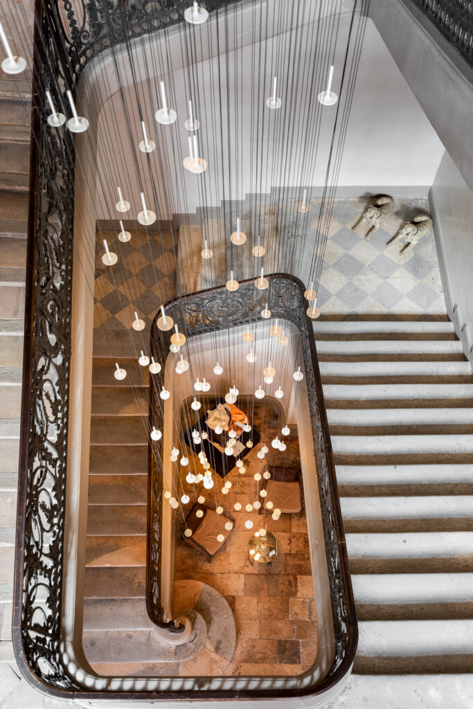 Vue plongeante sur les volées de l’escalier, les rampes ouvragées et les suspensions descendant jusqu’au hall.