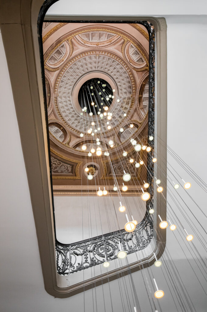 Perspective ascendante dans la cage d’escalier, entre un palier en ferronnerie, les suspensions et le plafond peint.