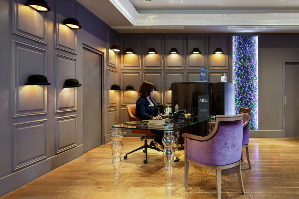 Réceptionniste derrière un bureau en verre, fauteuil violet et appliques en forme de chapeau à La Cour des Consuls.